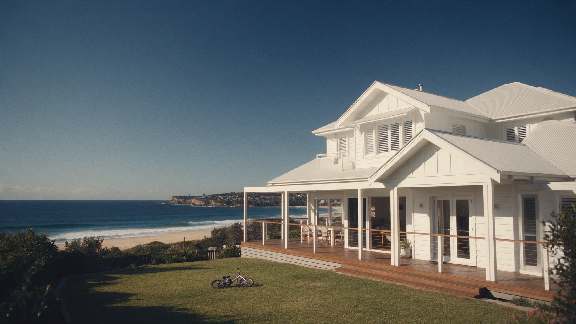 A white coastal family home with a timber deck and lawn.